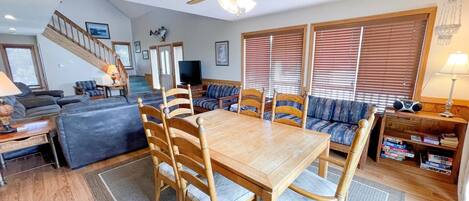 A spacious living room with a wooden dining table, comfortable seating, a ceiling fan, and a staircase leading to an upper level.