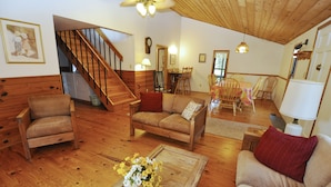 A wooden-floored living room with a sofa, chairs, a coffee table with flowers, a staircase, and a dining area. - Canel Cottage:  Tucked in the trees, nearby kayak launch area. (Ocracoke)