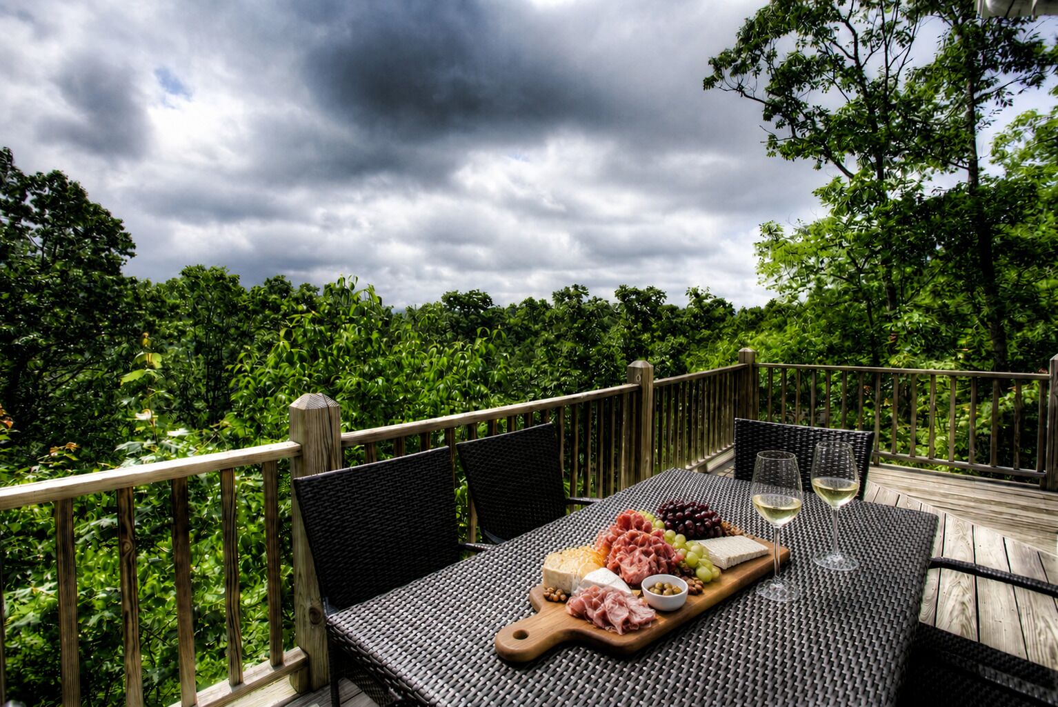 Un espacio de comedor al aire libre con sillas de mimbre y una mesa dispuesta con queso, embutidos, uvas y copas de vino.