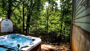 A hot tub surrounded by a wooden deck and dense forest, with a sign indicating spa rules.