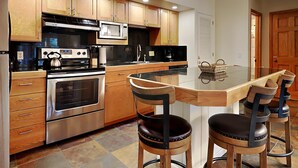 A modern kitchen with wooden cabinets, stainless steel appliances, and a glass dining table with bar stools. - 🌲Timberline C1C Standard 1-Bedroom ~Hot Tub~Pool~WiFi~Ski-in/out~Local Shuttle🌲 (Snowmass Village)
