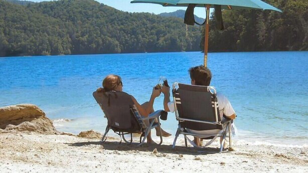 Two people relaxing on beach chairs under a beach umbrella, overlooking a lake with a forested hill in the background.