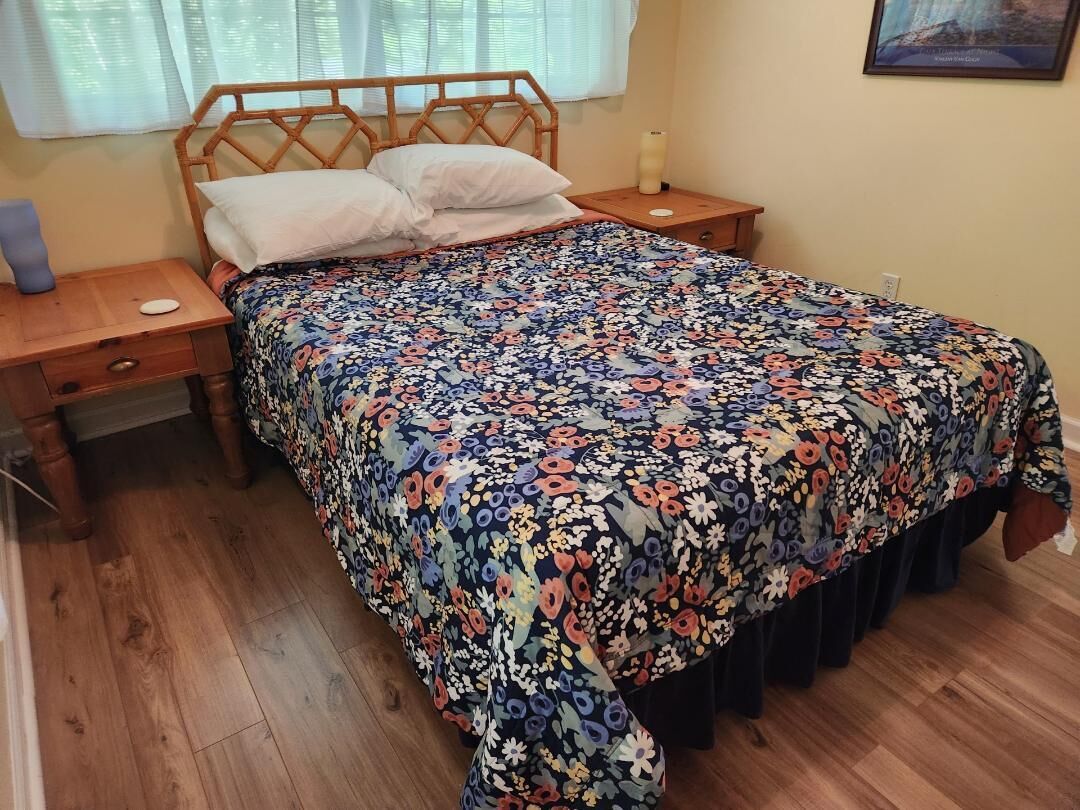 A bedroom with a wooden bed frame, floral bedspread, wooden nightstands, and a window with curtains.