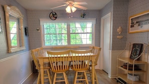 A dining area with a wooden table and chairs, a window with a view of greenery, a ceiling fan, and a shelf with items.
