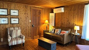 A cozy room with wooden walls, a striped armchair, a sofa with multiple cushions, a small table with a lamp, and framed fish pictures on the wall.