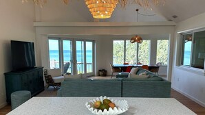 Amplia sala de estar con un gran ventanal con vista al océano, televisor y comedor con mesa y sillas.