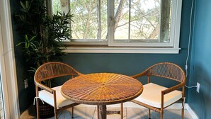 Una mesa redonda de mimbre con dos sillas, una lámpara colgante y una ventana con vistas a árboles.