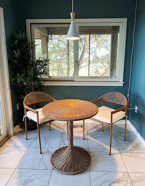A round wicker table with two chairs, a hanging light fixture, and a window with a view of trees.