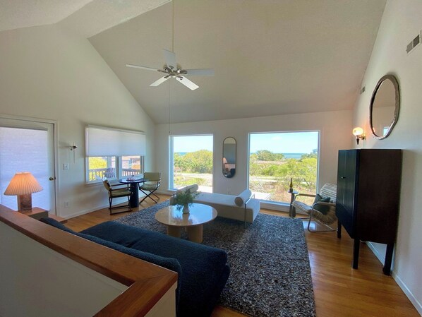 A spacious living room with a ceiling fan, large windows, a sofa, a coffee table, and a TV stand.