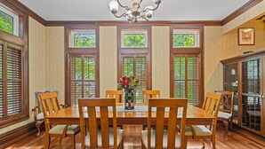 A dining room with a wooden table and chairs, large windows with shutters, and a chandelier.