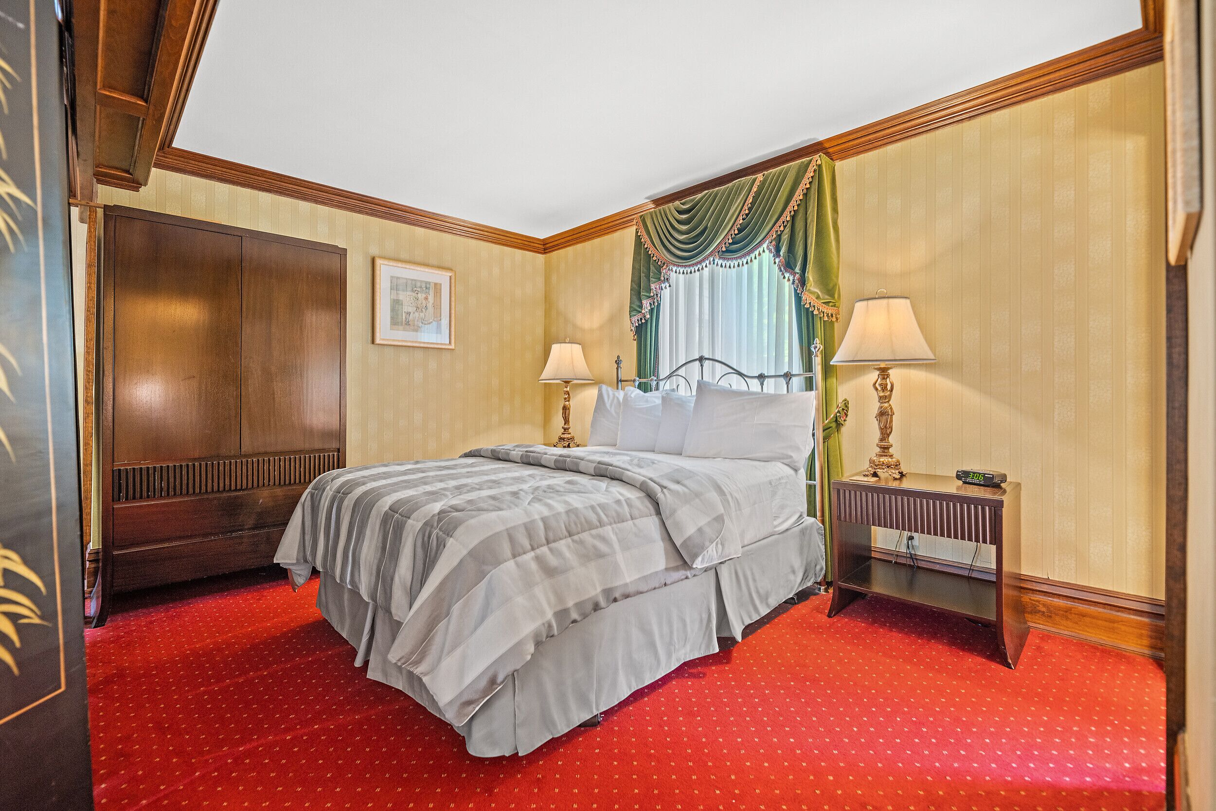 Een hotelkamer met een groot bed, twee nachtkastjes met lampen, een houten garderobe en een rood tapijt.
