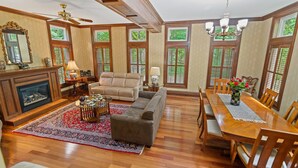 A spacious living room with hardwood floors, a fireplace, a sofa, a dining table with chairs, and large windows with shutters.