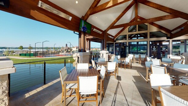 An outdoor patio with wooden tables and chairs, overlooking a body of water and a sports field. - Get Busy Livin! Grand Lake Waterfront (Afton)