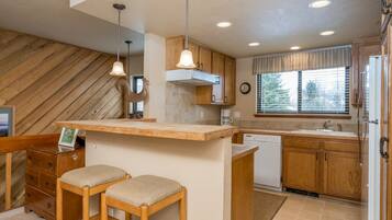 A compact kitchen with wooden cabinets, a built-in island with stools, a dishwasher, and a window with curtains.
