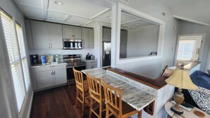 A compact kitchen with grey cabinets, a microwave, an oven, and a refrigerator. There is a wooden dining table with four chairs. - Daydreamer:  Reverse floorplan home offering three master bedrooms. (Ocracoke)