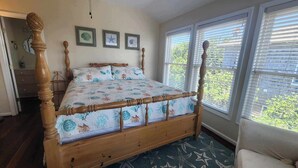 A wooden four-poster bed with a floral bedspread, framed pictures on the wall, and a view of greenery through the window blinds. - Daydreamer:  Reverse floorplan home offering three master bedrooms. (Ocracoke)