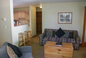 A living room with a sofa, a coffee table, and a kitchen area with wooden cabinets and stools.