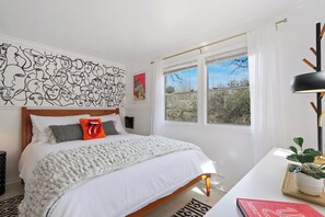 Un dormitorio con una cama grande, un cuadro con rostros, una ventana con vistas y una mesita de noche con una planta.