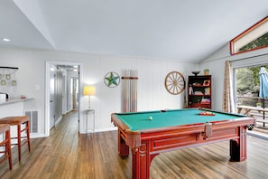 Una mesa de billar en una habitación con suelo de madera, una lámpara y una estantería.