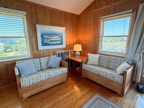 A cabin interior with wooden walls, two sofas with striped cushions, a table with a lamp, and a framed picture of a boat. - Ford Cottage: Beautiful marsh views with a large screened porch. (Ocracoke)