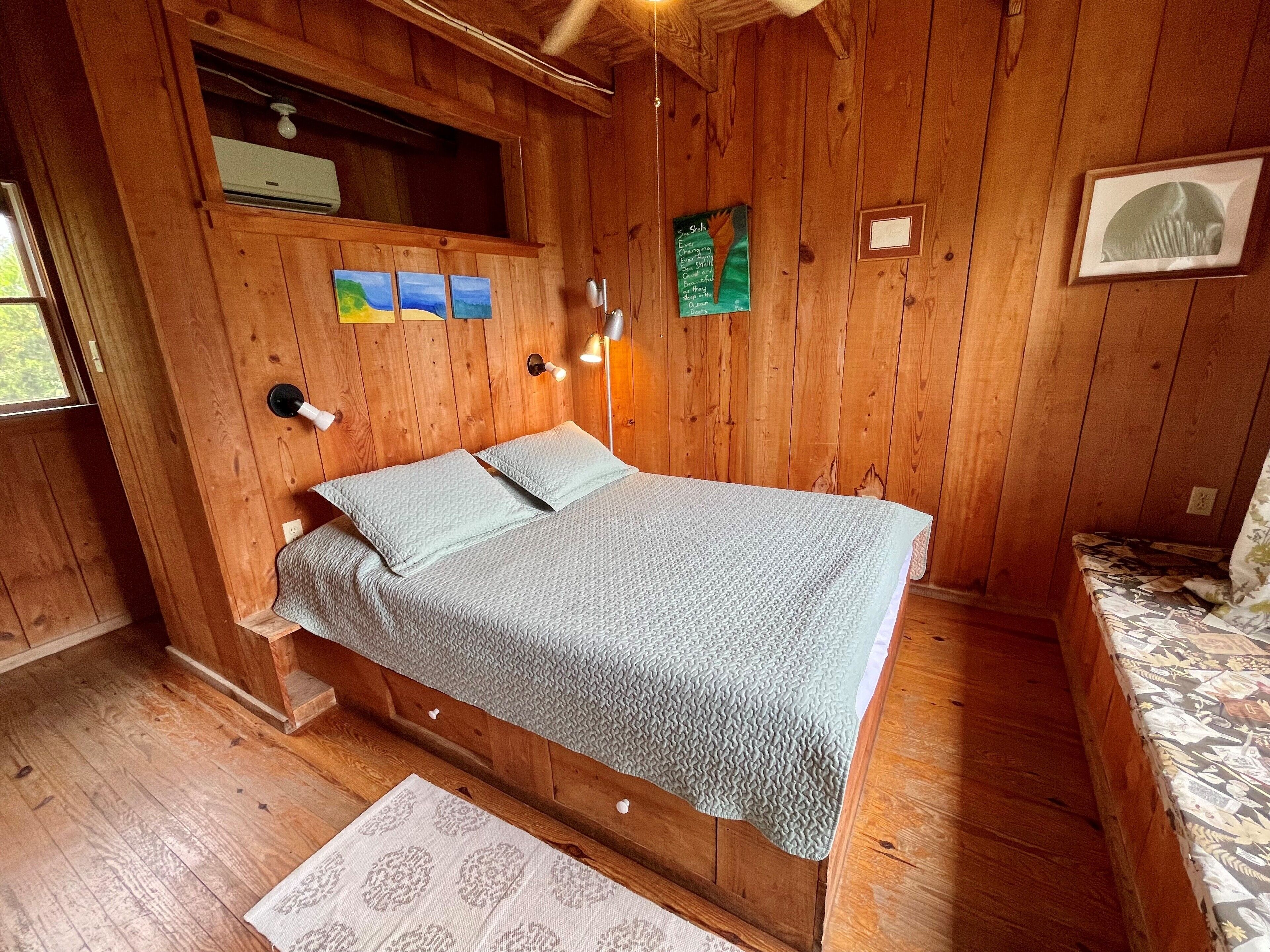 Una habitación de cabaña de madera con una cama, un estante pequeño con un calefactor y una ventana.