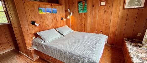 Una habitación de cabaña de madera con una cama, un estante pequeño con un calefactor y una ventana.
