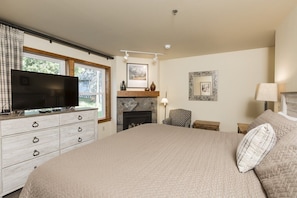 A bedroom with a large bed, a flat-screen TV on a dresser, a fireplace, and a window with curtains.