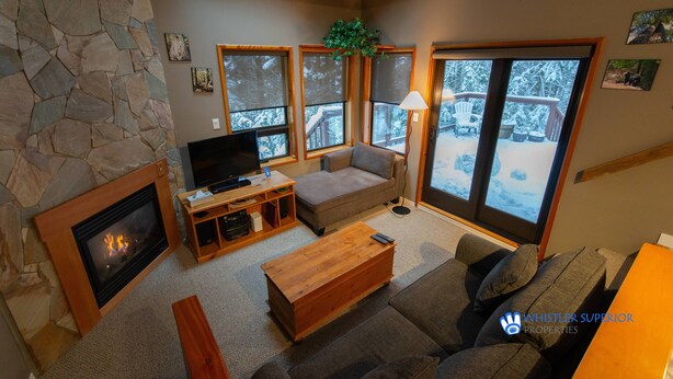 A cozy living room with a stone fireplace, comfortable seating, a television, and large windows offering a view of a snowy deck. - Cozy, Quiet Mountainside Location, Free Parking & WiFi, Huge Deck (Whistler)