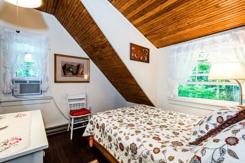 A bedroom with a sloped ceiling, a bed with floral bedding, a chair, a table, and a window with a view of greenery.