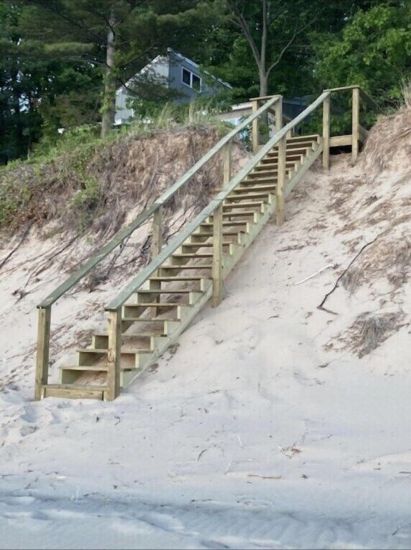 Una escalera de madera que sube por una pendiente arenosa, rodeada de árboles y una casa al fondo.