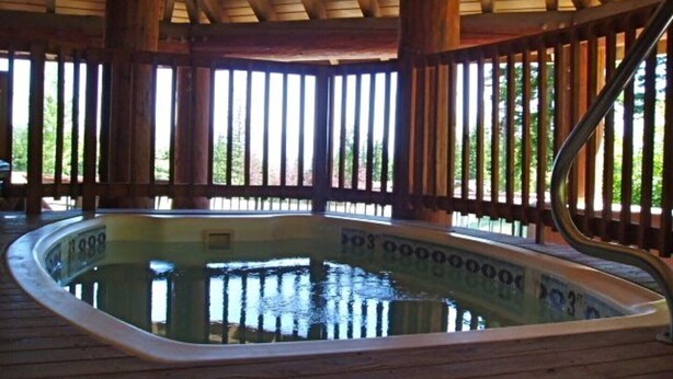 A hot tub with a wooden deck and railings, surrounded by a wooden structure with a sloped roof. - Charming Condo with Lots of Room (Whitfish)