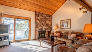 A cozy living room with a stone fireplace, a sofa, a television, and a large window overlooking a deck. - Sunburst Condo 2749 - Comfortable Elkhorn Space Off of Golf Course w/ Mt View (Elkhorn)