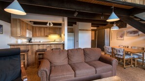 Ein gemütliches Wohnzimmer mit braunem Sofa, einem Holzespeisetisch und Stühlen. Es gibt eine Küchenzeile mit Holzschränken und einer Mikrowelle.