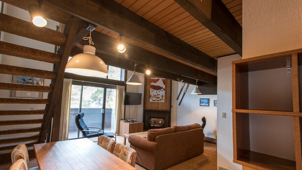 Ein gemütliches Wohnzimmer mit Holztisch, Sofa und Kamin. Hängelampen und ein Fernseher sind an der Wand angebracht.