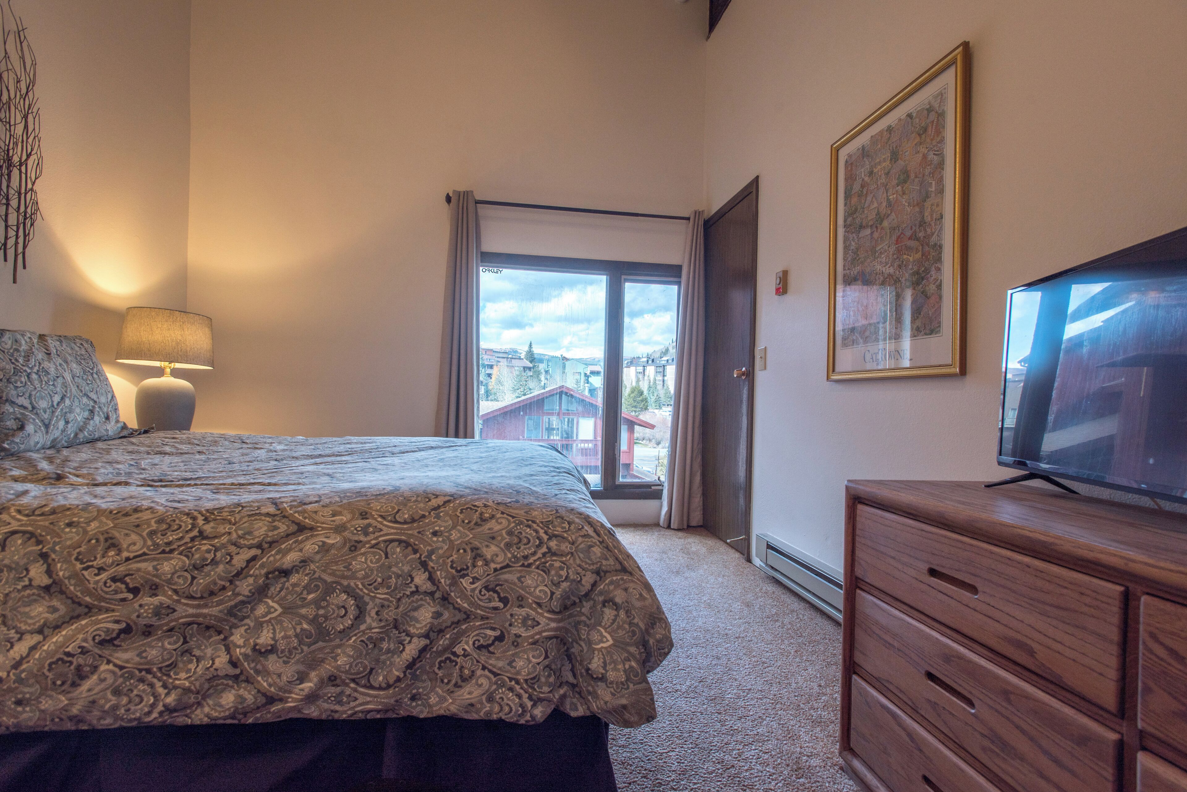 Ein Schlafzimmer mit Bett, Kommode, Fernseher, Fenster und Blick auf Gebäude sowie einer Tür.