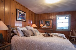 Une chambre à coucher aux murs lambrissés, avec un lit, deux lampes, un tableau encadré, une fenêtre et une chaise.