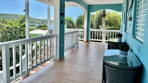 A covered balcony with a view of a lake and hills, white railings, and a small table with a plant.