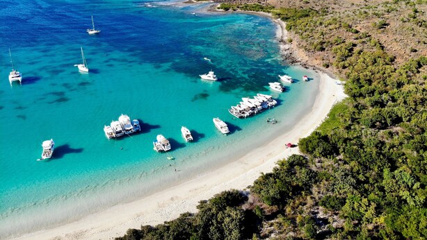 An aerial view of a coastal area with boats docked at a sandy beach, surrounded by greenery and rocky outcrops.