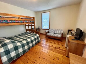 A small, cozy room with a bunk bed, a striped armchair, a desk with a computer, and a window with blinds.