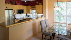 A modern kitchen with a stainless steel refrigerator, microwave, and coffee maker. There is a glass dining table with chairs and a window with blinds. - Alpine Greens - Village Shuttle, BBQ, Patio, Free Parking & WiFi (Whistler)