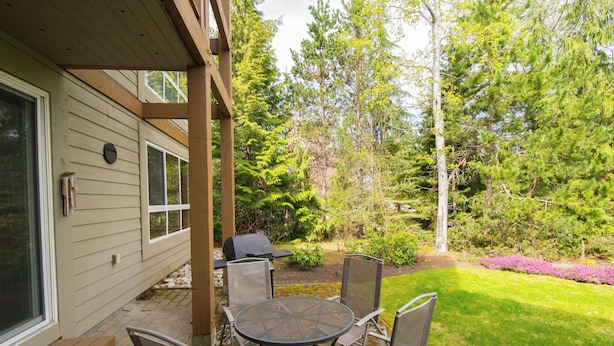 An outdoor patio with a round table and chairs, a grill, and a view of a forested area. - Alpine Greens - Village Shuttle, BBQ, Patio, Free Parking & WiFi (Whistler)