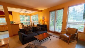 A living room with a sofa, coffee table, and a dining table with chairs. There are large windows with blinds, a kitchen area, and a ceiling fan. - Alpine Greens - Village Shuttle, BBQ, Patio, Free Parking & WiFi (Whistler)