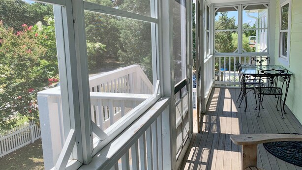 A covered porch with a wooden deck, a table with chairs, and a view of a garden with trees and flowers. - Sun Kissed: Cheerfully decorated dog friendly cottage. (Ocracoke)