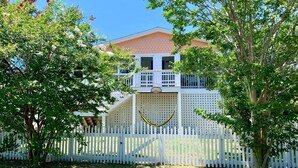 A two-story house with a balcony, surrounded by a white picket fence and lush greenery. - Sun Kissed: Cheerfully decorated dog friendly cottage. (Ocracoke)