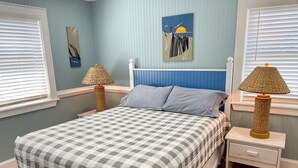 A bedroom with a checkered bedspread, two bedside tables with lamps, a nightstand, and a wall painting. - Sun Kissed: Cheerfully decorated dog friendly cottage. (Ocracoke)
