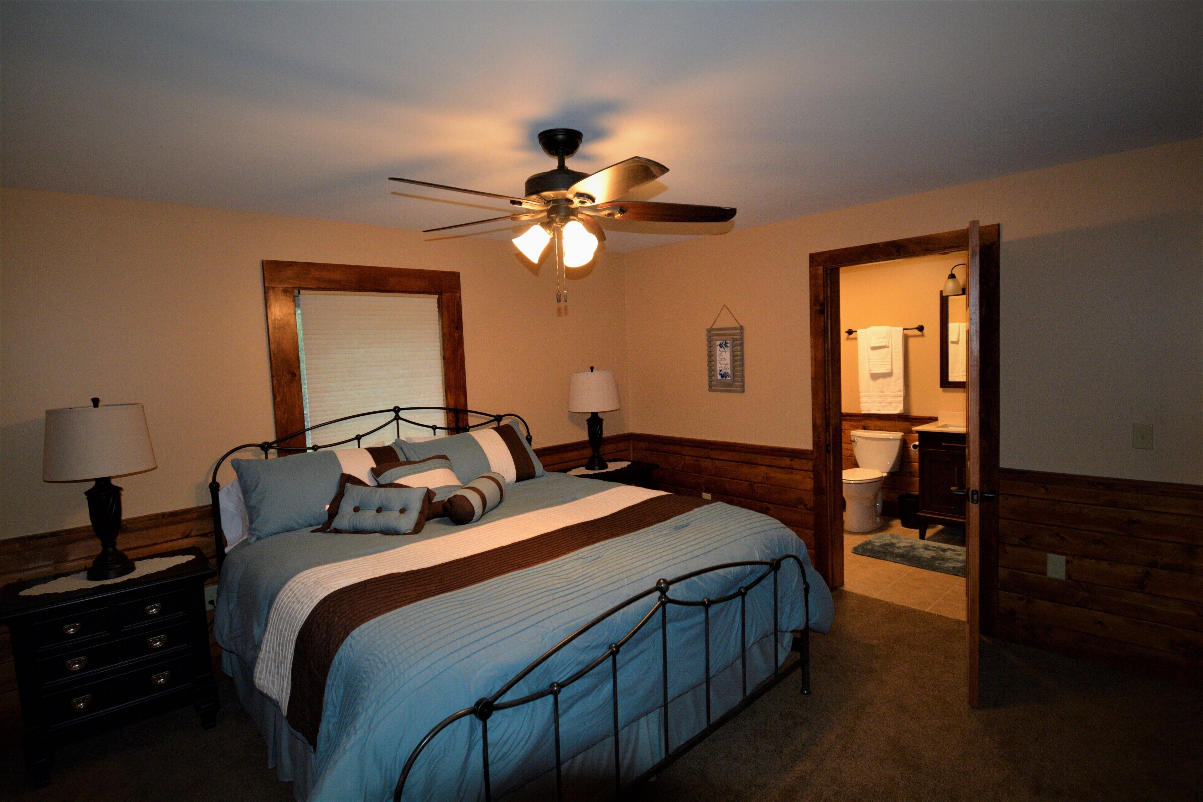 A bedroom with a bed, two bedside tables, a ceiling fan, a window with blinds, and a door leading to a bathroom.