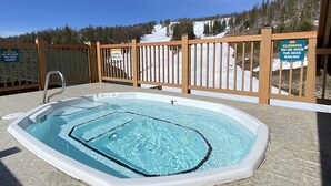 Een hottub met een houten terras en leuning, omringd door een besneeuwd landschap en bomen.
