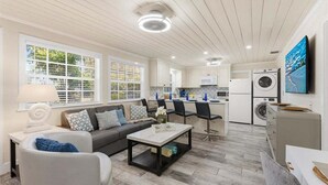 Una cocina moderna con armarios blancos, un microondas y una lavadora. Hay una zona de comedor con una mesa y sillas, y una sala de estar con un sofá y una mesita de centro.