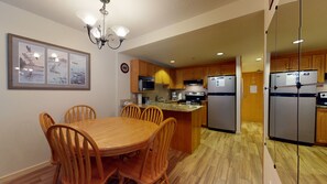 A kitchen with wooden cabinets, a refrigerator, a microwave, and a dining area with a table and chairs. - Woodrun Lodge - True Ski In/Ski Out, Pool, Hot Tub, Perfect for Families (Whistler)
