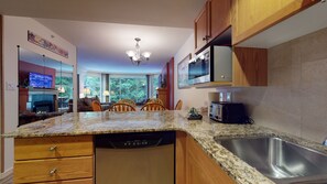 Una cocina con encimeras de granito, armarios de madera, lavavajillas integrado, pileta de acero inoxidable y un comedor con vista a árboles.